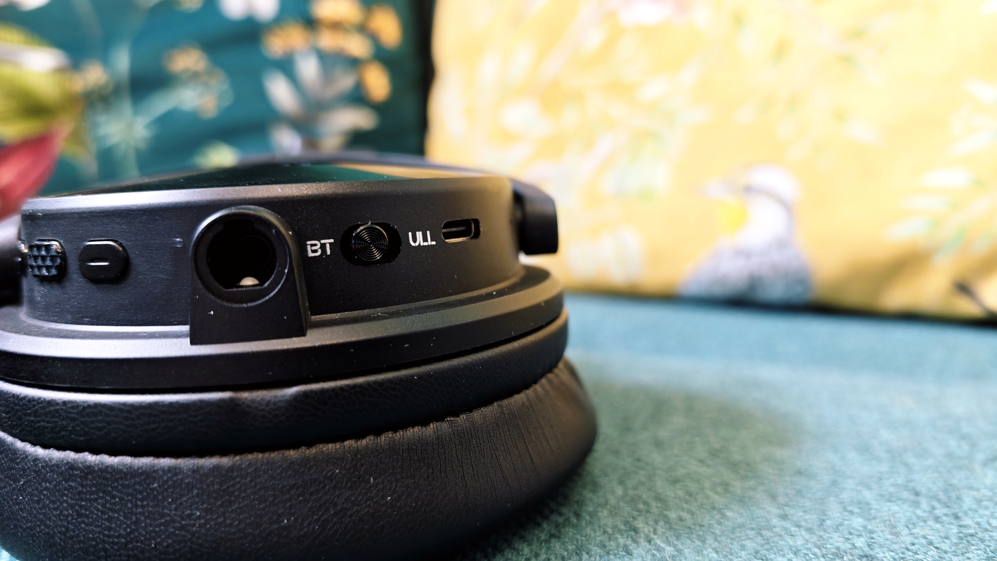 A pair of black OneOdio Studio Max 2 headphones with red internal earcups sitting on a green sofa in front of colourful pillows with birds and flowers on them
