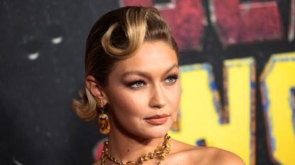 Gigi Hadid with a curled fringe wearing gold necklaces, in front of a black background with red and yellow writing