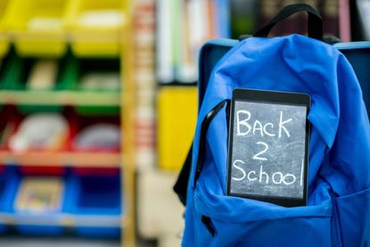 A tablet computer is sitting on a backpack The backpack is hanging from a classroom chair The tablet has back 2 school written on it