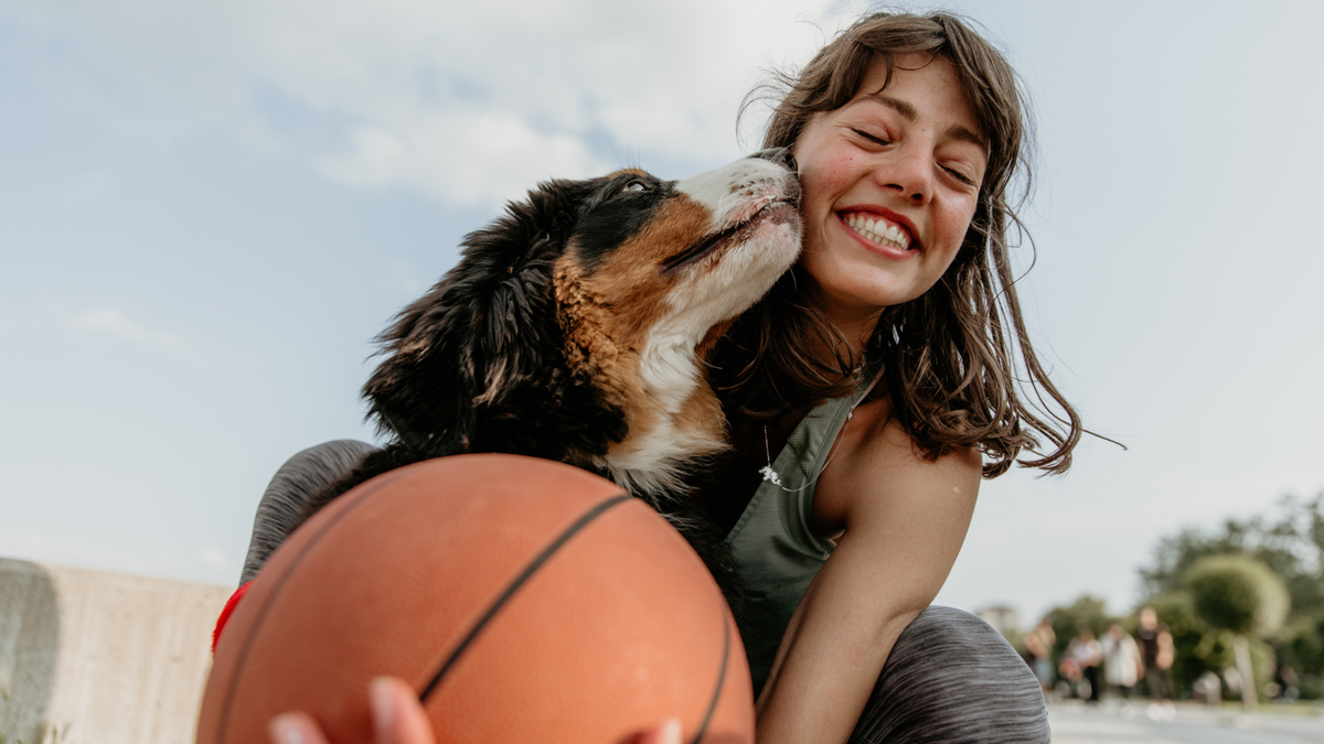 Woman laughing as a dog licks her face and she holds a basket ball