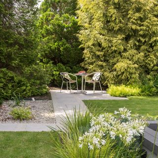 a path leading to a bistro set on a small compact patio area with flowers and planting all around