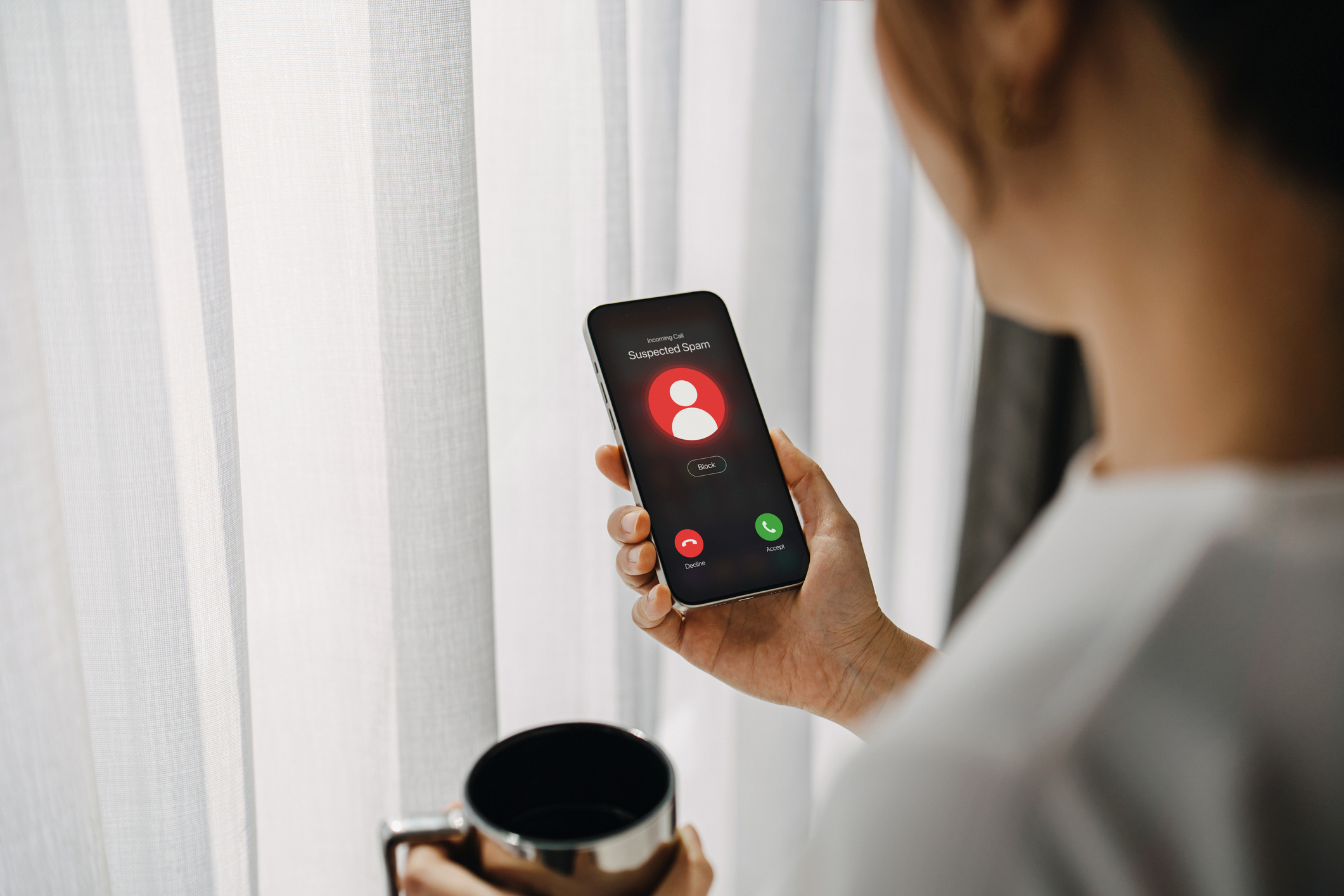 Young Asian woman receiving an incoming suspected call from unknown caller on her smartphone and rejecting the call at home. Device screen showing suspected scam as detected by network provider. 