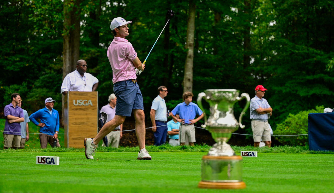 Evan Beck strikes a tee shot in front of the US Mid Amateur Championship trophy