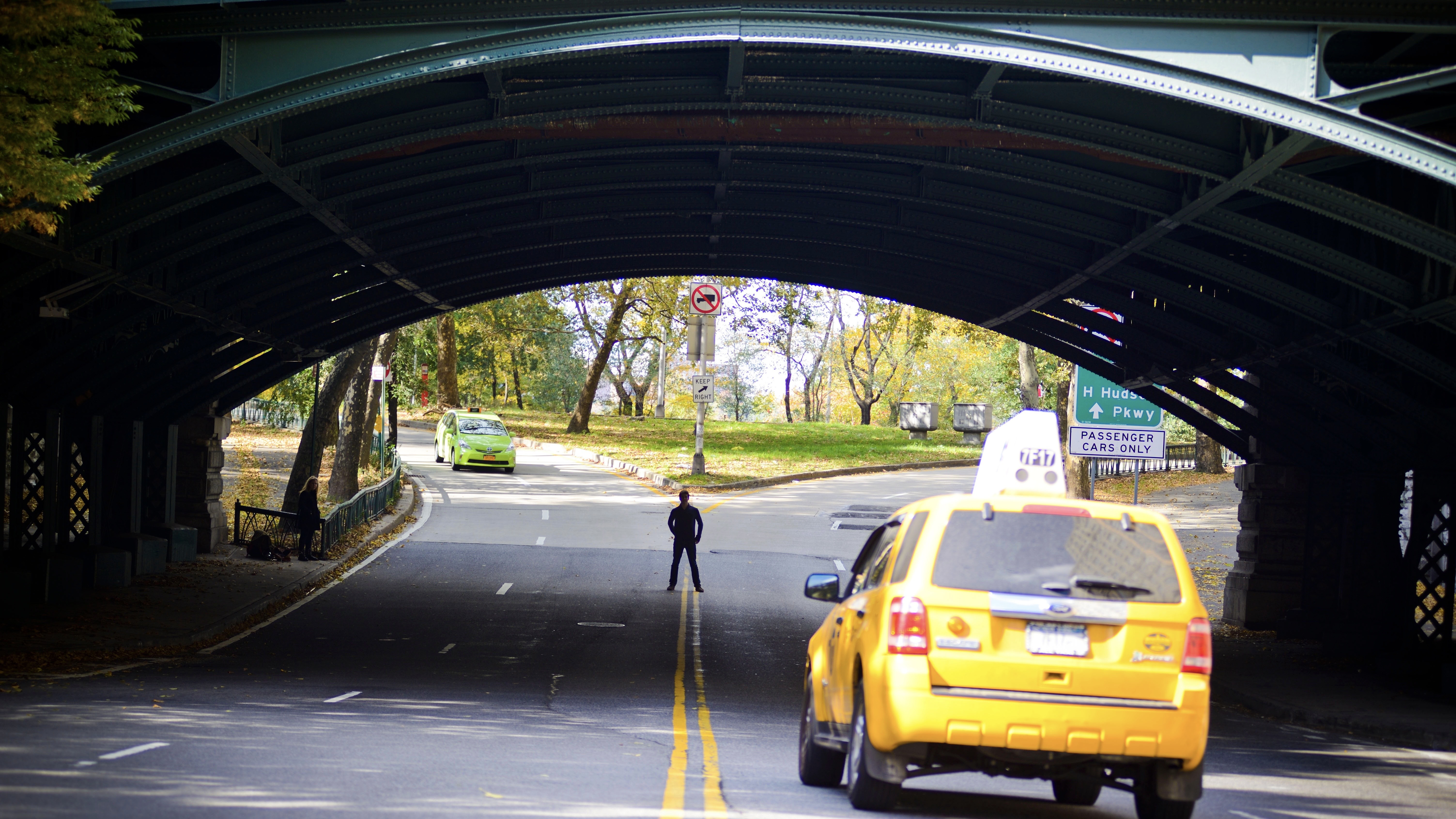 A small silhouette sands in the distance, in the middle of the road, past a green overpass. A taxi heads his direction in the foreground.