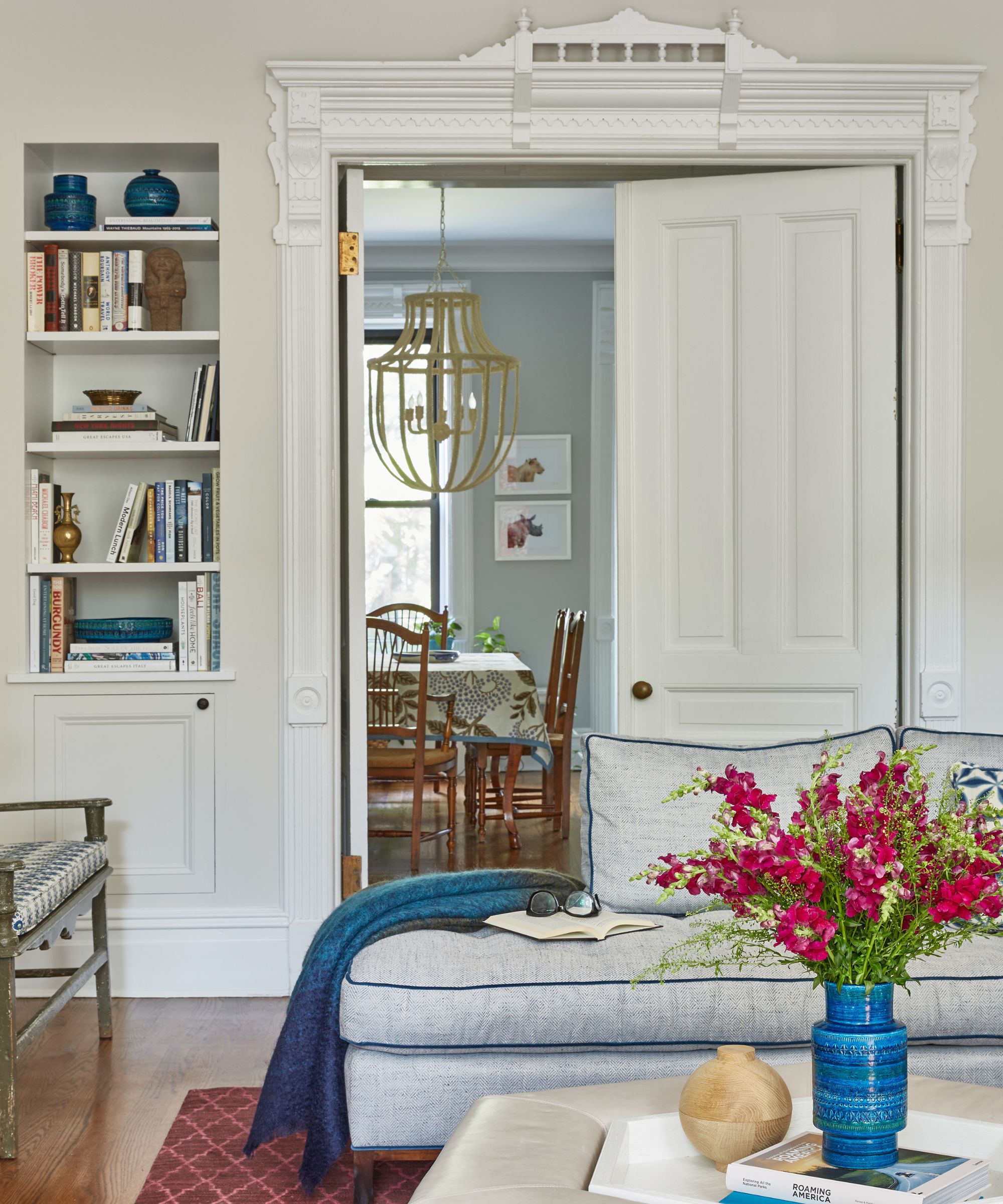 Loft living room with piped sofa, coffee table with blue vase, built-in bookshelves in an alcove, a red rug and large double doors looking into the dining room