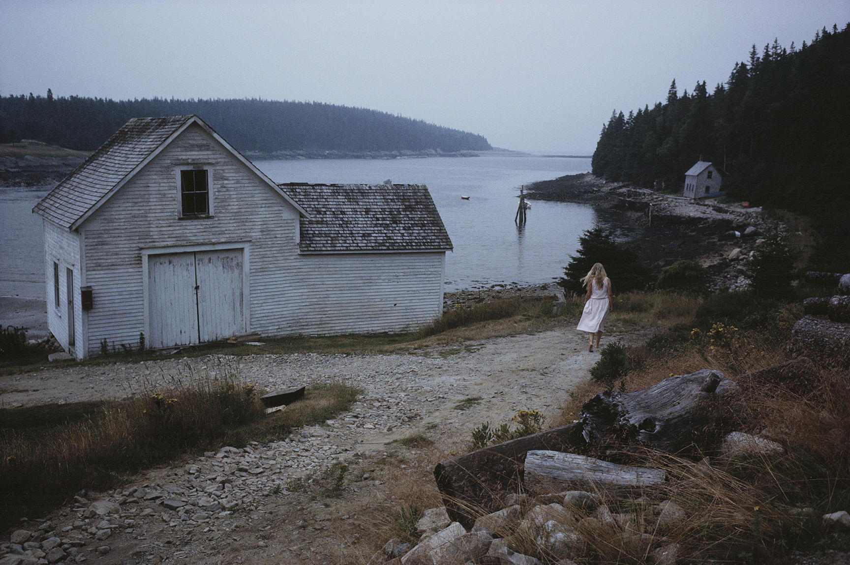 an old house on the lake front with a leading path and an individual in a white dress standing on it