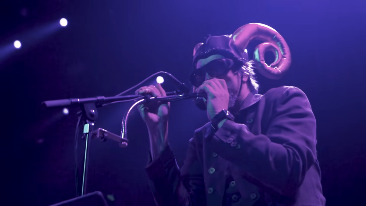 A picture of Les Claypool singing live on stage and wearing a ram-horned helmet