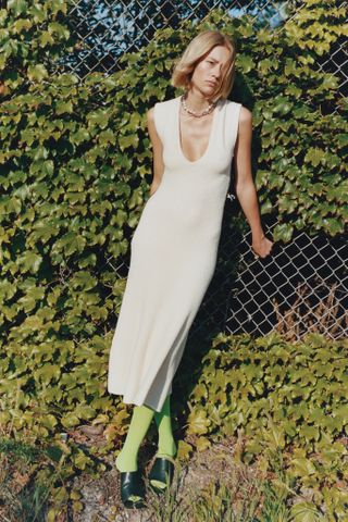 A model in a white Maria McManus dress posing in front of ivy and a chain fence