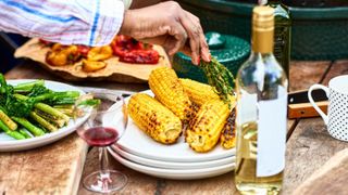 Grilled corn on a plate at a party with a glass of wine, a bottle of wine, being brushed with rosemary