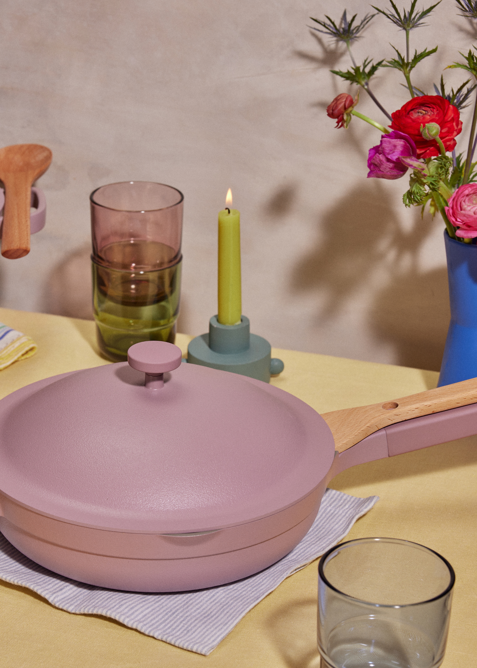 A dining table set with dark rose-colored ceramic pans, beside colored stacked glasses, a floral arrangement in a blue vase and a green candle