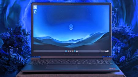 An Alienware 15 laptop sits on a cloth surface with its display showing a blue landscape wallpaper and the Alienware head logo. The background behind the laptop features glowing blue sci‑fi scenery.