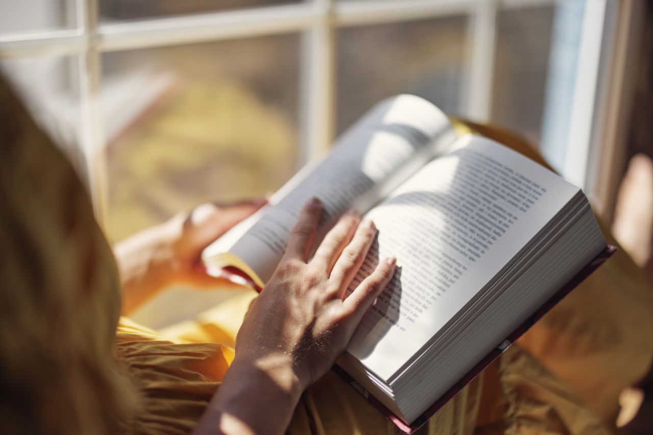 Photo over a person&#039;s shoulder as they read a book by a window