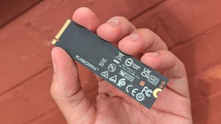 A hand holds a SanDisk SSD against a reddish-brown wooden background. The SSD features various certification logos, giving a tech-focused, modern feel.