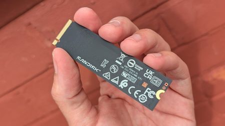A hand holds a SanDisk SSD against a reddish-brown wooden background. The SSD features various certification logos, giving a tech-focused, modern feel.