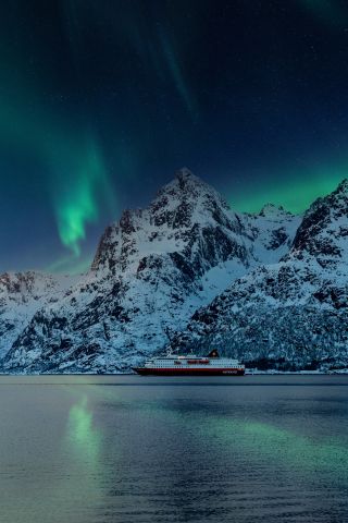 hurtigruten ship under the northern lights with tall mountains in the background.