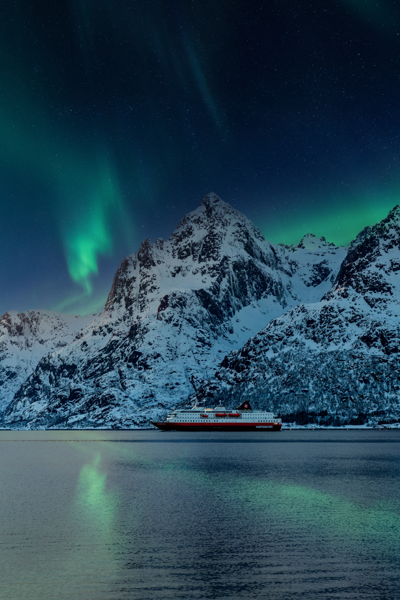 hurtigruten ship under the northern lights with tall mountains in the background.