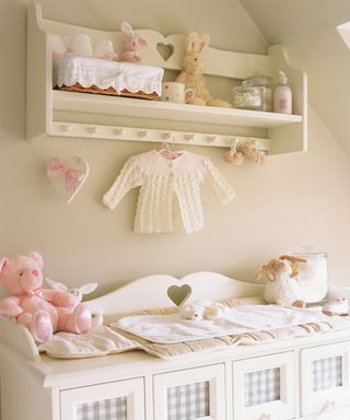 A baby girl nursery idea with white walls, white changing table, white shelf and pink toys and accessories.