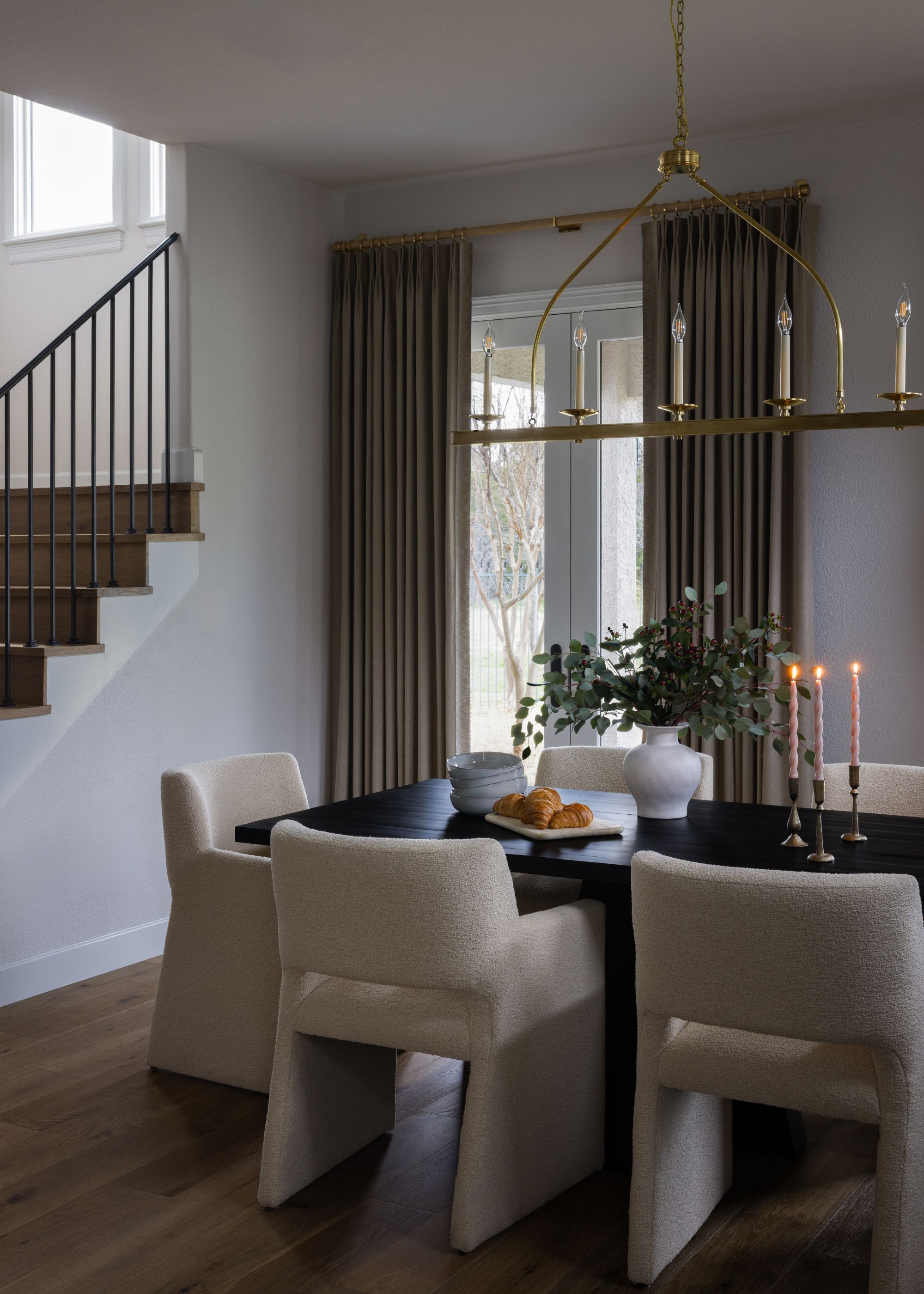 A modern dining area with a dark wood table surrounded by six cream-colored upholstered chairs. A large brass linear chandelier hangs above, and heavy taupe drapes frame the patio doors.
