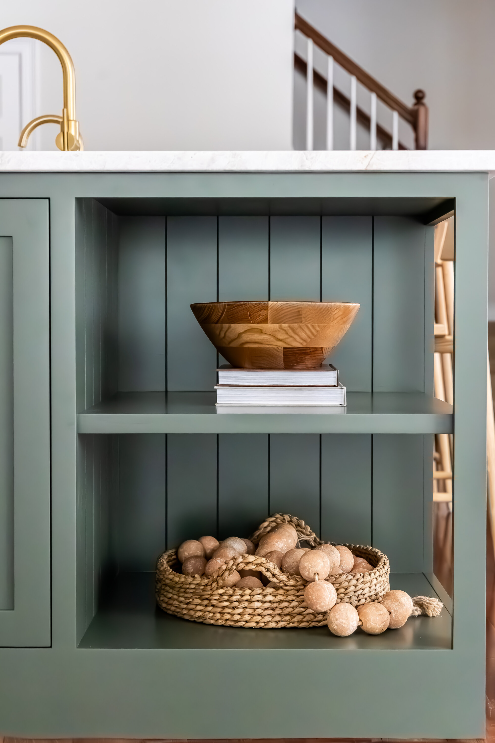 Close-up image of a shelf built into a kitchen island. It's painted with a dark moss green paint. On the shelves there is a wooden bowl and a woven basket with wooden decor.