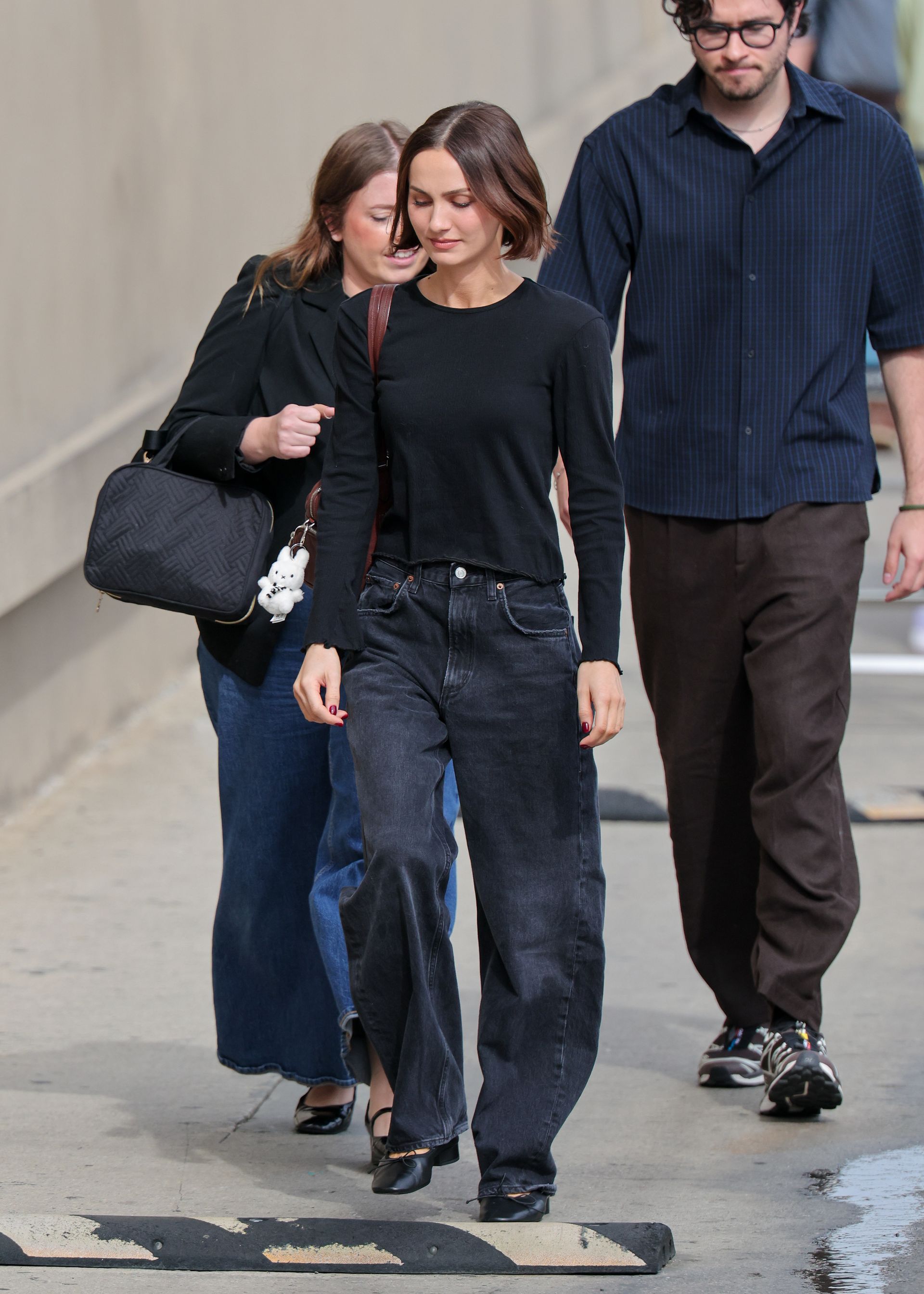 Maude Aptow wears black baggy jeans with a black longsleeve top and black heeled ballet pumps.