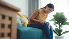 Asian woman suffering from knee pain, massage her knee by hands and sitting on sofa in living room at home.