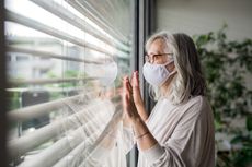 Woman with face mask standing indoors by window at home.