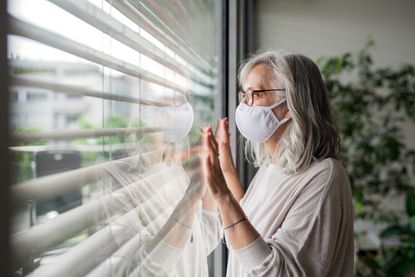 Woman with face mask standing indoors by window at home.