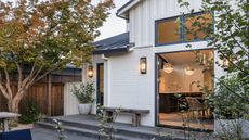 a backyard of a home with trees and plants placed all around. The exterior of the home is black and grey and you can see inside the bright and chic home.