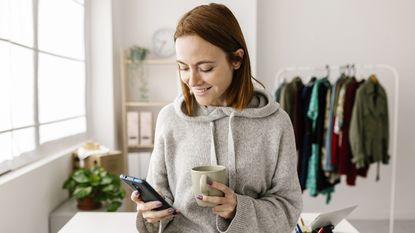 Smiling young adult woman looks at her smartphone while also holding a cup of coffee. 