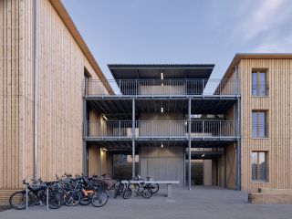 Refugee housing by Stocker Dewes Architekten. A view between two blocks of a housing project with walkways between them, wooden walls and many bicycles parked in front of it.