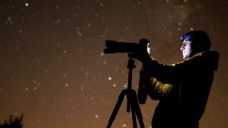 A man looking through a tripod-mounted camera with a long lens attached to it with a starry night sky in the background. 