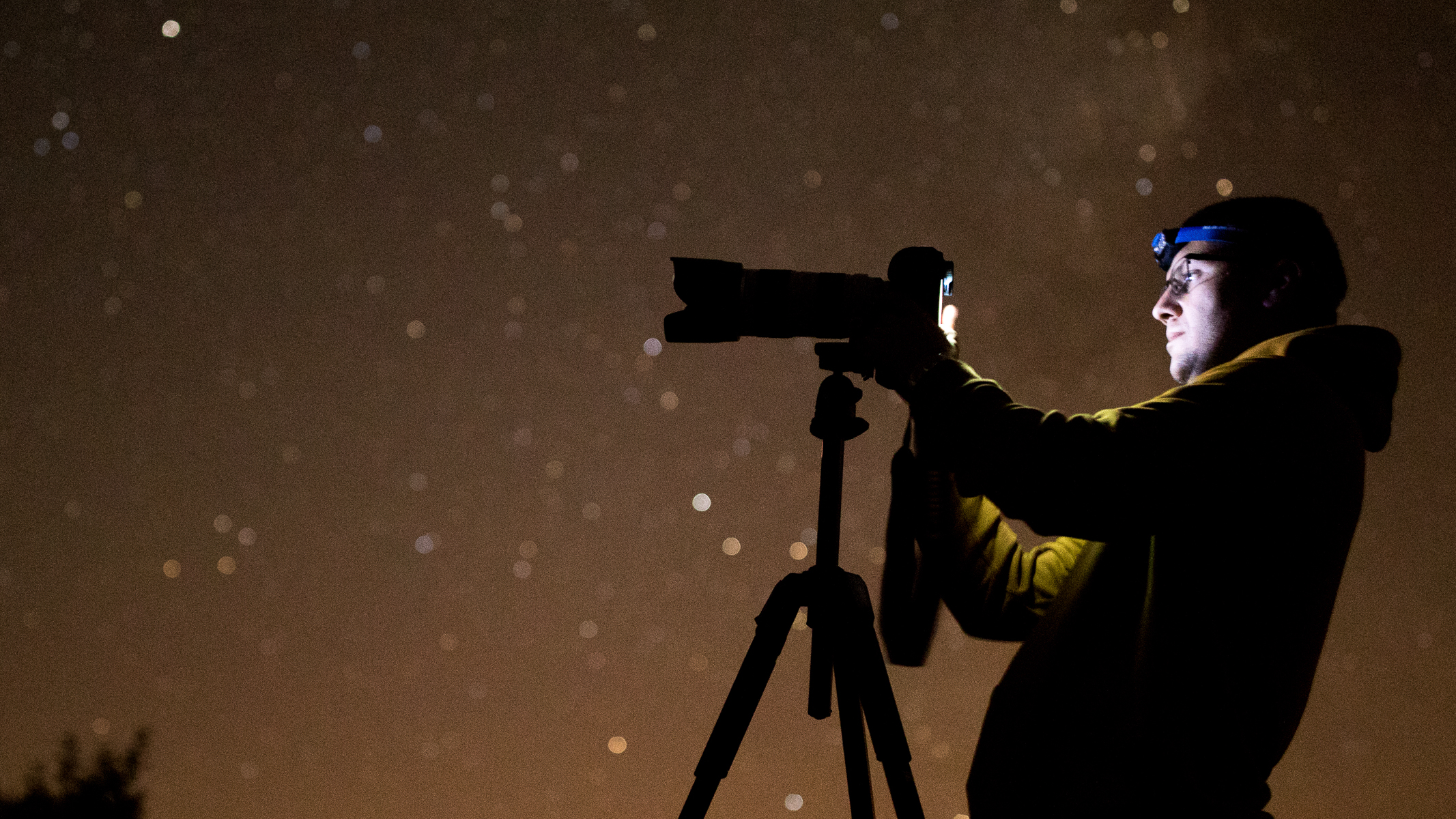A man looking through a tripod-mounted camera with a long lens attached to it with a starry night sky in the background. 