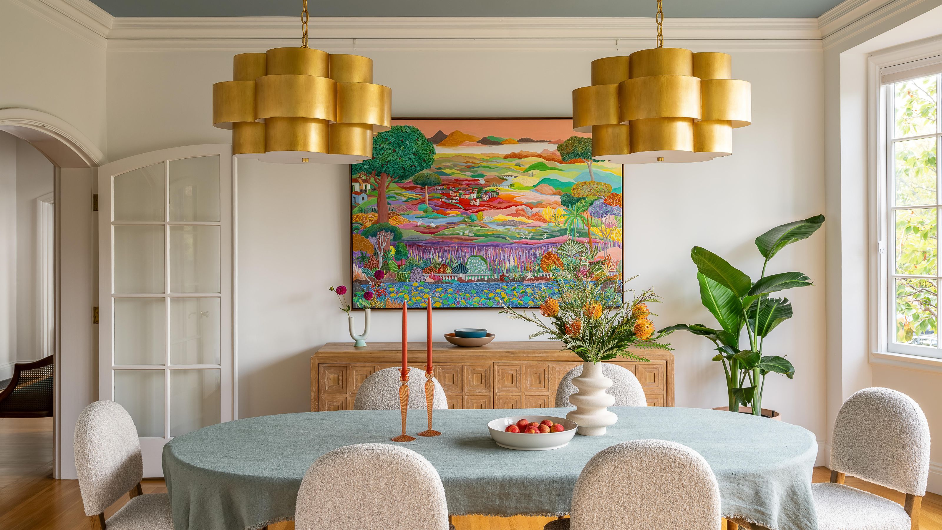 a white dining room with large arched doors and windows are either end, A blue painted accent ceiling and two brass statement pendants that hang over the table