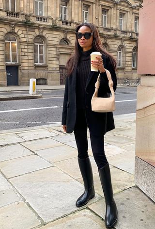 best riding boots shown in a photo of a woman standing outside wearing a boxy black blazer over a black turtleneck with black skinny jeans and black knee-high boots