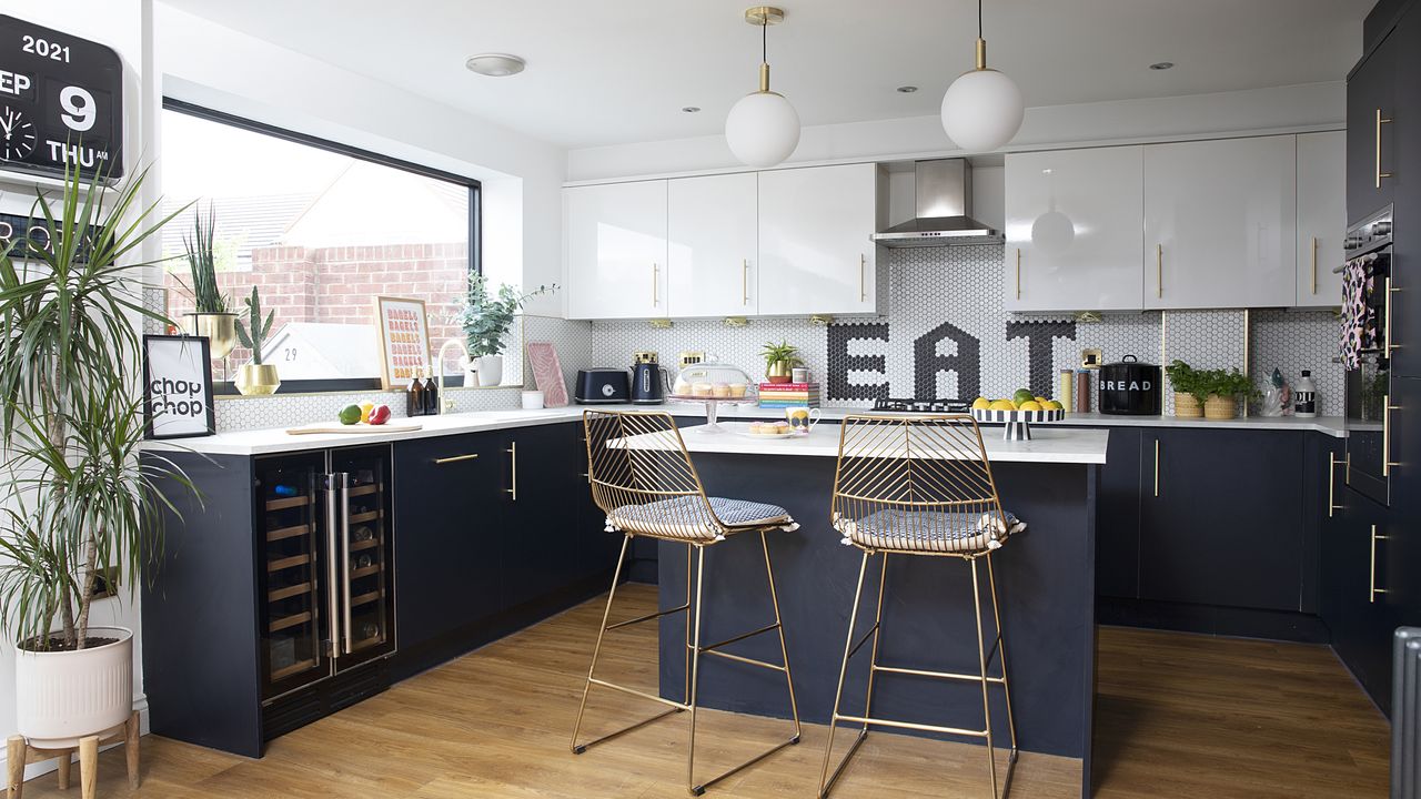 Blue U-shaped kitchen and island with white wall units, brass bar stools and white honeycomb splashback with black tiles forming the word &#039;eat&#039;