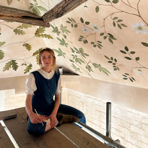 Image of a woman in denim dungarees sitting on a wooden structure with a painted ceiling above her.