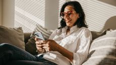 Woman sitting on sofa at home, wearing shirt, jeans, and glasses, smiling and scrolling on iPhone to represent the Paired app review