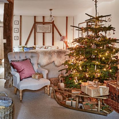 a living room with Christmas tree with twinkling fairy lights and an ornamental sledge covered in wrapped Christmas presents beside a traditional armchair