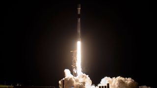 a white and black rocket lifts into the predawn sky, lighting its launch pad and ground below