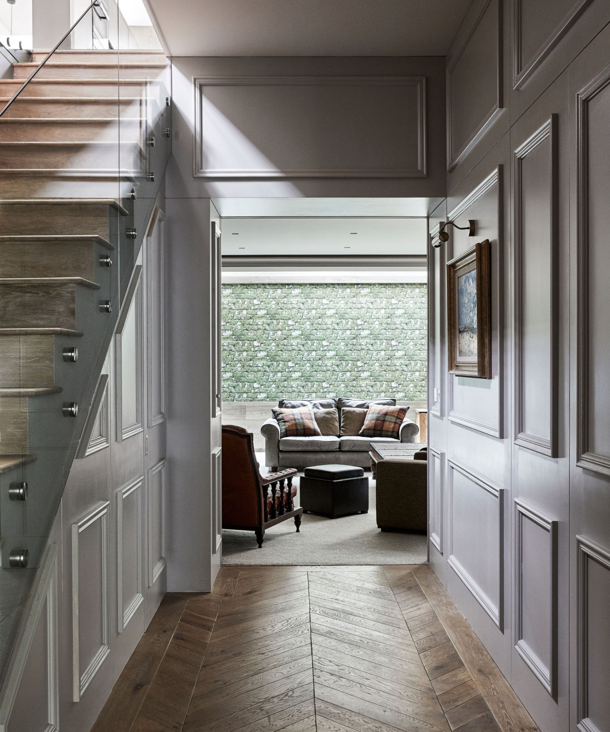 An entryway space with neutral walls, wooden flooring, wooden stairs with a glass balustrade, and a living room with green walls seen beyond.