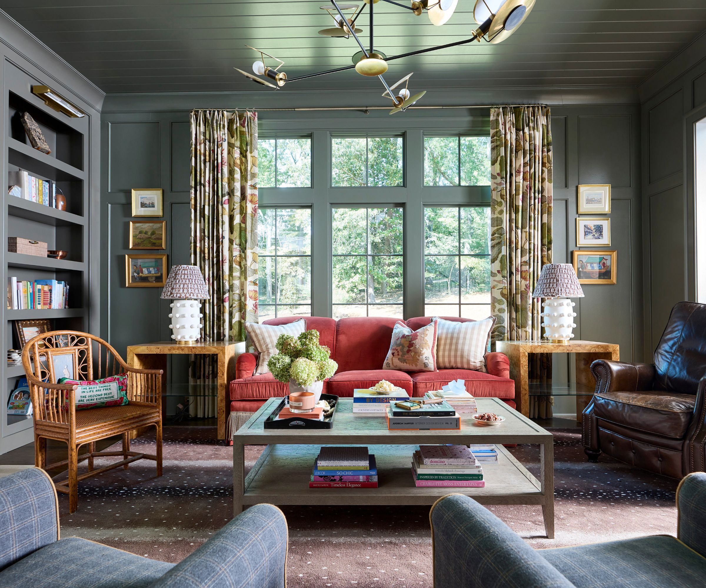 A living room with muted green color-drenched walls and ceiling, built-ins, a raspberry red sofa in front of the windows, and traditional decor.