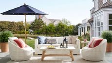 An outdoor dining scene with white rattan seats, a coffee table with lanterns on it, a blue parasol, and houses in the background