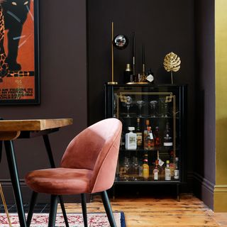 a dark painted dining room with a pink upholstered dining chair and gold drinks cabinet with gold stripe on the wall