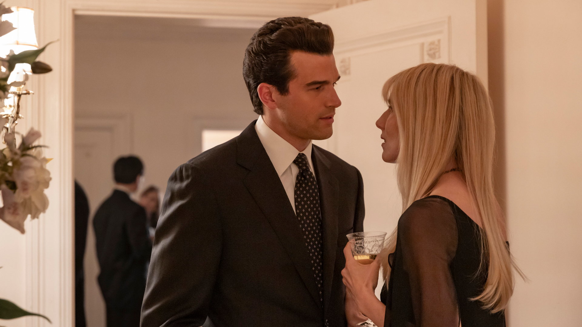 A man in a suit (Paul Anthony Kelly as JFK Jr.) stands with a blonde woman in a black top (Dree Hemingway as Daryl Hannah), who holds a champagne glass. The pair stand next to a flower arrangement in a room of a grand house, in a still from 'FX's Love Story: John F. Kennedy Jr. &amp; Carolyn Bessette.'