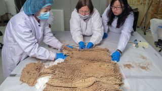 Three women in lab coats lean over a piece of armor made in a fish scale design