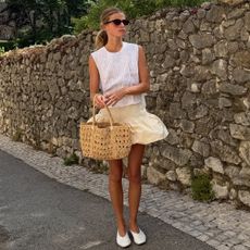 A woman wears a white top, peplum skirt, raffia bag, and white ballet flats.