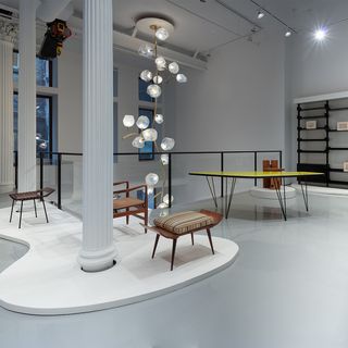 Inside R & Company’s new gallery space featuring three brown stalls of different styles, including one with a multi-colour stripe cushion. Also featuring a floor-to-ceiling flower-like light and a yellow-top table.