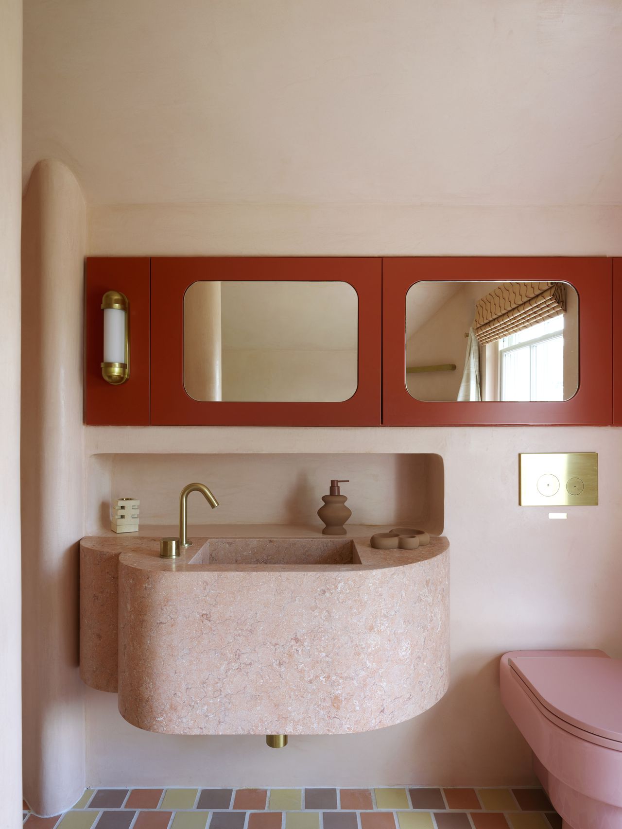 Image of a light pink bathroom with a matching pink terrazzo sink. There is a row of red cabinets above the sink that have mirrors on the doors. The toilet is pink.