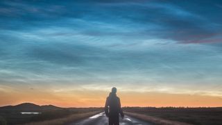 A man stannds on a road with shining silver clouds in the dark sky above