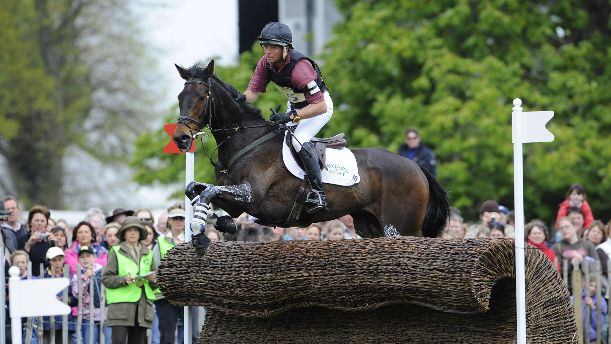 Dan Jocelyn, a rider in a dark red top with a black body protector, rides dark bay horse Silence over a a fence shaped like a wave at Badminton Horse Trials